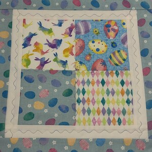 K&ouml;nnte beinhalten: Ein quadratischer Quiltblock zum Thema Ostern mit vier Stoffquadraten. Das obere linke Quadrat zeigt bunte Hasen, das obere rechte Ostereier, das untere linke Ostereier und das untere rechte ein Rautenmuster. Der Block ist mit wei&szlig;em Stoff und einer wellenf&ouml;rmigen Naht eingefasst.