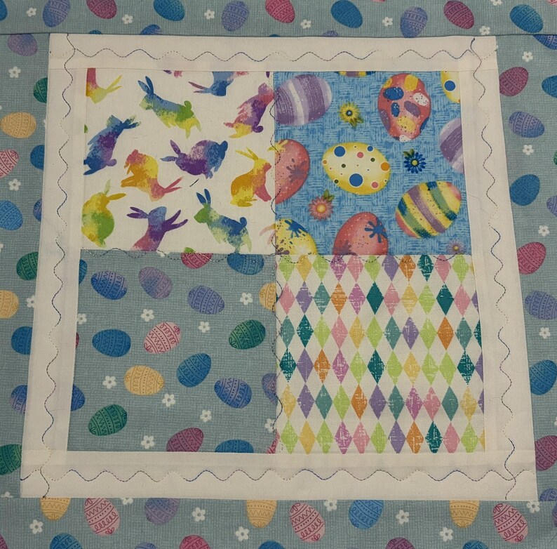 K&ouml;nnte beinhalten: Ein quadratischer Quiltblock zum Thema Ostern mit vier Stoffquadraten. Das obere linke Quadrat zeigt bunte Hasen, das obere rechte Ostereier, das untere linke Ostereier und das untere rechte ein Rautenmuster. Der Block ist mit wei&szlig;em Stoff und einer wellenf&ouml;rmigen Naht eingefasst.