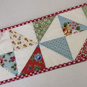 May include: A colorful quilted table runner featuring a patchwork design with alternating white triangles and patterned fabric triangles. The fabrics include floral and polka dot patterns in red, blue, green, and yellow. The runner is trimmed with a red and white gingham border.