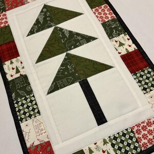May include: A quilted Christmas table runner featuring three green Christmas trees with white outlines on a white background. The trees have a black trunk. The border is made of red, white, and green patterned fabric.