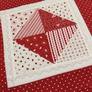 May include: A quilted placemat with a central diamond design composed of red and white fabrics. The diamond features heart and stripe patterns, with a scalloped edge and a red polka dot border. A Valentine's Day theme.