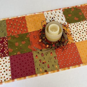 May include: A rectangular patchwork table runner featuring a variety of autumn-themed fabric squares in shades of orange, green, burgundy, and cream. The runner is topped with a glass candle holder containing a white pillar candle and a berry wreath.