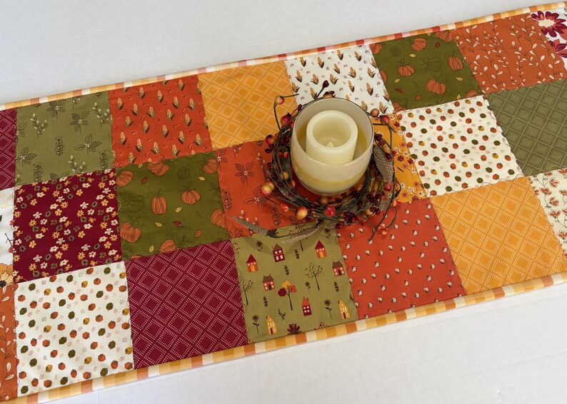 May include: A rectangular patchwork table runner featuring a variety of autumn-themed fabric squares in shades of orange, green, burgundy, and cream. The runner is topped with a glass candle holder containing a white pillar candle and a berry wreath.