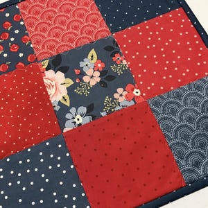 May include: A patchwork square featuring nine fabric squares in red, navy, and floral patterns. The red squares have white polka dots or are solid. Navy squares have white dots, apples, or a fan design. The edges are finished with a navy blue border.