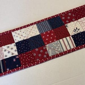 May include: A rectangular quilted table runner featuring a patchwork design of red, white, and navy blue fabric squares. The pattern includes stars, stripes, floral motifs, and text listing various U.S. states. It is finished with a red fabric border.