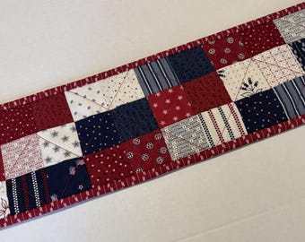 May include: A rectangular quilted table runner featuring a patchwork design of red, white, and navy blue fabric squares. The pattern includes stars, stripes, floral motifs, and text listing various U.S. states. It is finished with a red fabric border.
