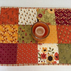 Peut inclure: Un tapis en tissu patchwork rectangulaire compos&eacute; de douze carr&eacute;s aux motifs automnaux vari&eacute;s dans des tons orange, jaune, vert et bordeaux. Un bougeoir en verre orange avec une bougie pilier blanche est plac&eacute; au centre du tapis.