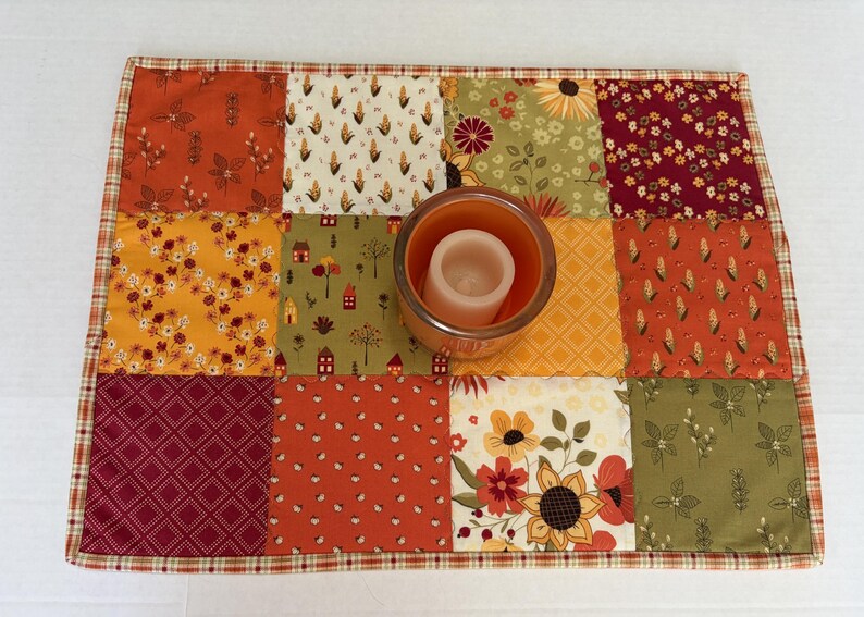 Peut inclure: Un tapis en tissu patchwork rectangulaire compos&eacute; de douze carr&eacute;s aux motifs automnaux vari&eacute;s dans des tons orange, jaune, vert et bordeaux. Un bougeoir en verre orange avec une bougie pilier blanche est plac&eacute; au centre du tapis.