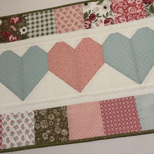 May include: A quilted table runner featuring a repeating pattern of pink and blue hearts with white polka dots. The runner is bordered by various floral and plaid fabric patches in shades of pink, green, and white. The runner is approximately 12 inches wide.