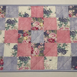 May include: A patchwork quilt with a floral pattern in shades of pink, white, and blue. The quilt is made up of squares and triangles, and the fabric is a mix of gingham and floral prints.