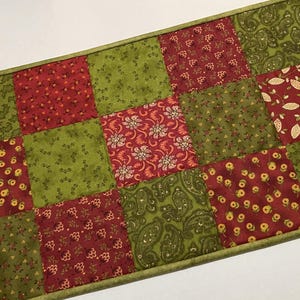May include: A quilted table runner featuring a patchwork design. The runner is composed of alternating squares of red and green fabric. The red squares have floral and leaf patterns, while the green squares have various patterns. The edges are trimmed with a green border.