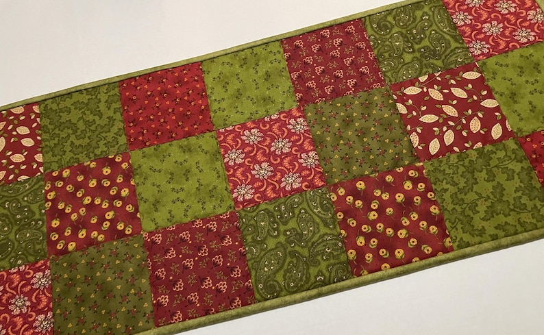 Puede incluir: Camino de mesa acolchado con dise&ntilde;o de patchwork. El camino de mesa est&aacute; compuesto por cuadrados alternos de tela roja y verde. Los cuadrados rojos tienen estampados florales y de hojas, mientras que los cuadrados verdes tienen varios estampados. Bordes verdes.