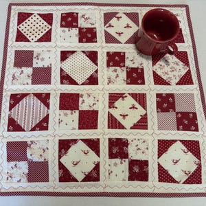 May include: A square fabric placemat featuring a patchwork design of sixteen blocks in shades of red and white. The pattern includes alternating squares of floral prints, polka dots, bee motifs, and bird illustrations, finished with a wavy cream border.