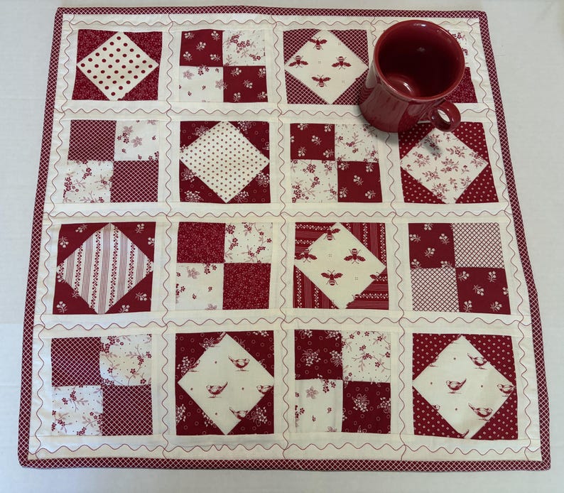 May include: A square fabric placemat featuring a patchwork design of sixteen blocks in shades of red and white. The pattern includes alternating squares of floral prints, polka dots, bee motifs, and bird illustrations, finished with a wavy cream border.