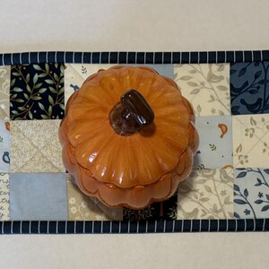 May include: A rectangular quilted table runner featuring a patchwork design of navy blue, light blue, and cream fabric squares with floral, bird, and script patterns. A glossy orange ceramic pumpkin-shaped lidded jar sits in the center.