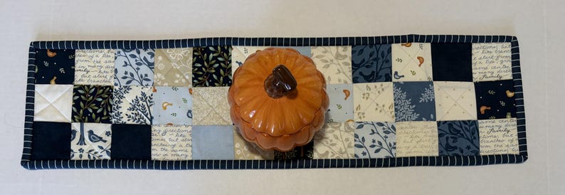 May include: A rectangular quilted table runner featuring a patchwork design of navy blue, light blue, and cream fabric squares with floral, bird, and script patterns. A glossy orange ceramic pumpkin-shaped lidded jar sits in the center.