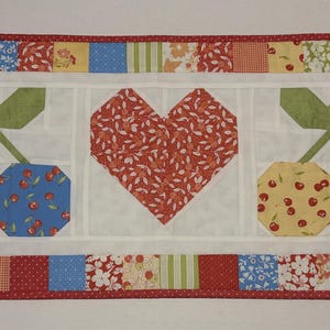 May include: A rectangular quilted table runner featuring a central design of a red heart and two pairs of cherries. The cherries are blue and yellow with red accents. The border is a patchwork of floral and patterned fabrics.