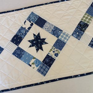 May include: A quilted table runner featuring a white background with a diamond-stitched pattern. The runner is decorated with repeating square blocks containing dark blue eight-pointed stars surrounded by various blue and white floral prints.