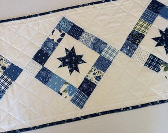 May include: A quilted table runner featuring a white background with a diamond-stitched pattern. The runner is decorated with repeating square blocks containing dark blue eight-pointed stars surrounded by various blue and white floral prints.