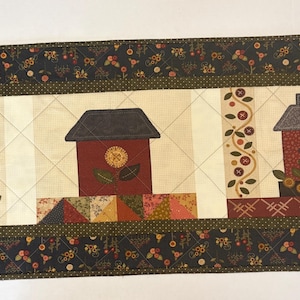 May include: A rectangular quilted table runner featuring a repeating pattern of houses in shades of red and dark gray, accented with floral motifs, a vine with circular shapes, and a border of dark fabric with a small floral print and cross-stitching.
