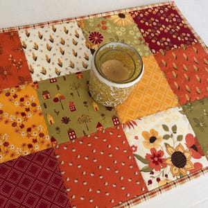 Peut inclure: Un tapis en tissu matelass&eacute; rectangulaire pr&eacute;sentant un motif patchwork aux th&egrave;mes automnaux dans des tons orange, jaune, rouge et vert olive, avec un bougeoir en verre plac&eacute; au centre contenant une bougie &agrave; la cire de couleur claire.