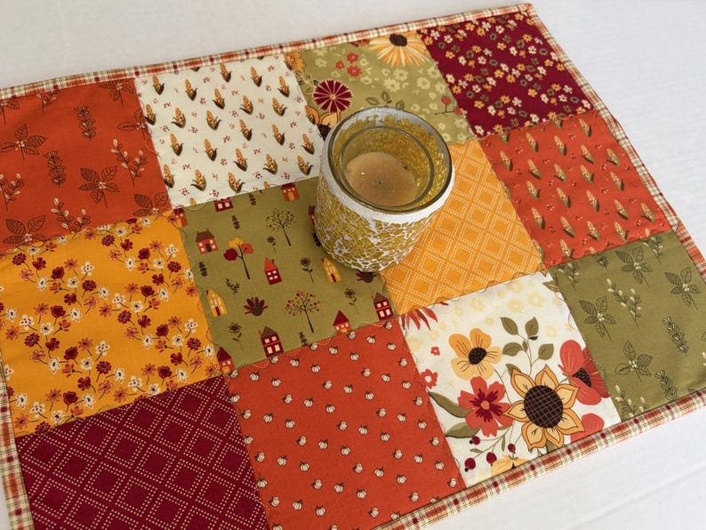Peut inclure: Un tapis en tissu matelass&eacute; rectangulaire pr&eacute;sentant un motif patchwork aux th&egrave;mes automnaux dans des tons orange, jaune, rouge et vert olive, avec un bougeoir en verre plac&eacute; au centre contenant une bougie &agrave; la cire de couleur claire.