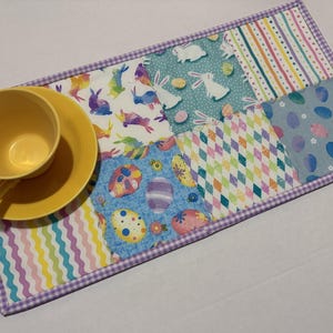 Może przedstawiać: Ż&oacute;łty filiżanka i spodek spoczywają na kolorowej podkładce w tematyce wielkanocnej. Podkładka ma wzory patchworkowe z kr&oacute;liczkami, jajkami i geometrycznymi wzorami, obramowanymi fioletowym wykończeniem w kratkę vichy. Projekt jest jasny i świąteczny.