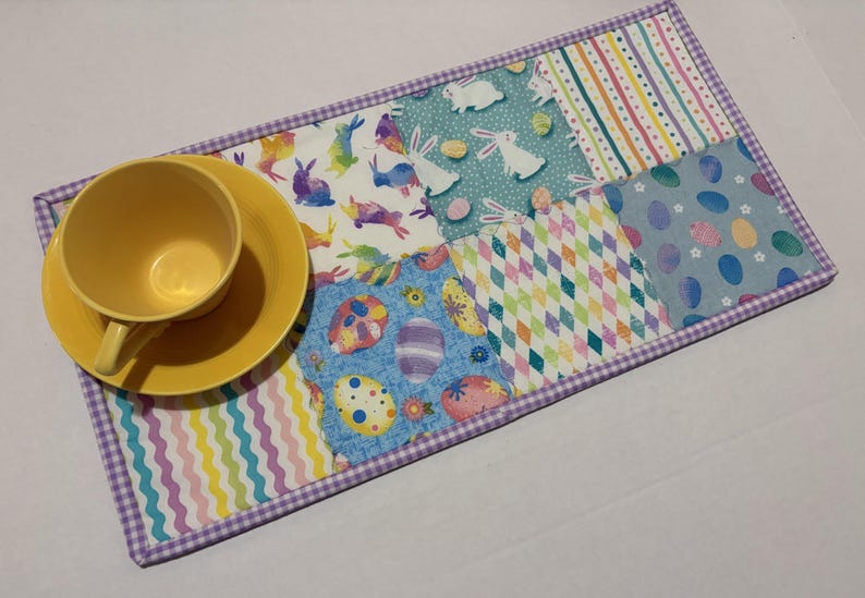 Może przedstawiać: Ż&oacute;łty filiżanka i spodek spoczywają na kolorowej podkładce w tematyce wielkanocnej. Podkładka ma wzory patchworkowe z kr&oacute;liczkami, jajkami i geometrycznymi wzorami, obramowanymi fioletowym wykończeniem w kratkę vichy. Projekt jest jasny i świąteczny.