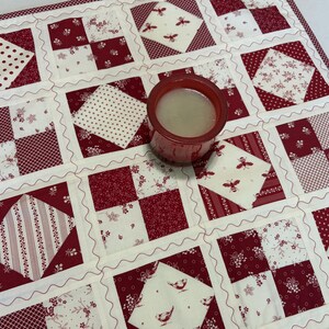 May include: A square quilted fabric mat featuring a patchwork design of red and white squares with various patterns including polka dots, floral prints, stripes, and bee motifs, with a red glass candle holder containing wax placed in the center.