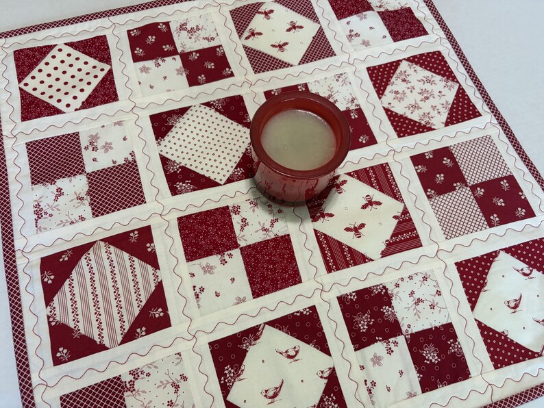 May include: A square quilted fabric mat featuring a patchwork design of red and white squares with various patterns including polka dots, floral prints, stripes, and bee motifs, with a red glass candle holder containing wax placed in the center.