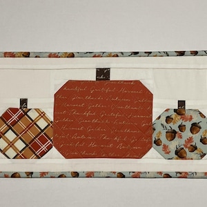 May include: A quilted table runner featuring five pumpkin shapes in various patterns and colors. The pumpkins include floral, plaid, and script designs, set against a white background. The runner is trimmed with a patterned border.