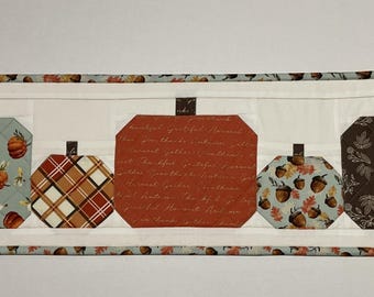 May include: A quilted table runner featuring five pumpkin shapes in various patterns and colors. The pumpkins include floral, plaid, and script designs, set against a white background. The runner is trimmed with a patterned border.