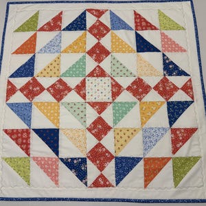 May include: A square quilt with a white background and a colorful patchwork design. The quilt features triangles and squares in red, blue, yellow, green, and orange floral patterns. A blue and white dotted border frames the quilt.
