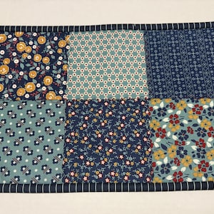 May include: A patchwork placemat with six fabric squares in various floral and geometric patterns. The colors include navy, teal, yellow, red, and white. The border is navy with white stripes.