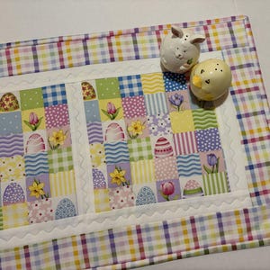 Pode incluir: Um caminho de mesa colorido com tema de P&aacute;scoa com um design de patchwork com ovos de P&aacute;scoa, flores e bolinhas. O caminho de mesa tem uma borda xadrez. Dois saleiros e pimenteiros de cer&acirc;mica, um coelhinho e um pintinho, est&atilde;o no caminho.