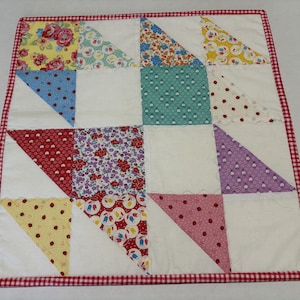 May include: A handmade patchwork quilt with a red and white gingham border. The quilt features a pattern of alternating white squares and colorful triangles with floral and polka dot designs. The quilt is approximately 20 inches square.