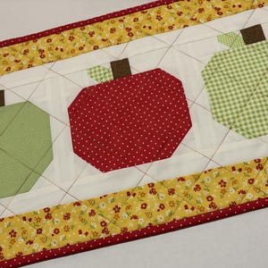 May include: A quilted table runner featuring three stylized apple shapes. The apples are green, red, and green gingham, with brown stems and green leaves. The runner has a white background with a yellow floral border and red trim.