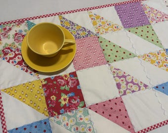 May include: A yellow ceramic teacup and saucer set placed on a rectangular quilted fabric mat. The mat features a patchwork design of various floral and patterned fabric triangles in shades of red, yellow, green, blue, and purple, with a red and white checkered border.