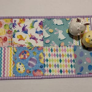 Może przedstawiać: Prostokątna podkładka na st&oacute;ł w tematyce wielkanocnej z patchworkowym wzorem przedstawiającym kolorowe wzory jajek, kr&oacute;lik&oacute;w i kształt&oacute;w geometrycznych. Dwie ceramiczne solniczki i pieprzniczki w kształcie kr&oacute;lika i kurczaka.