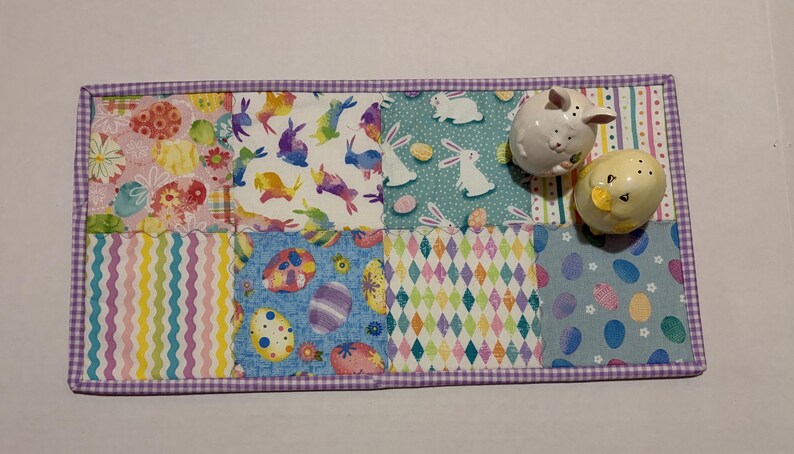 Może przedstawiać: Prostokątna podkładka na st&oacute;ł w tematyce wielkanocnej z patchworkowym wzorem przedstawiającym kolorowe wzory jajek, kr&oacute;lik&oacute;w i kształt&oacute;w geometrycznych. Dwie ceramiczne solniczki i pieprzniczki w kształcie kr&oacute;lika i kurczaka.