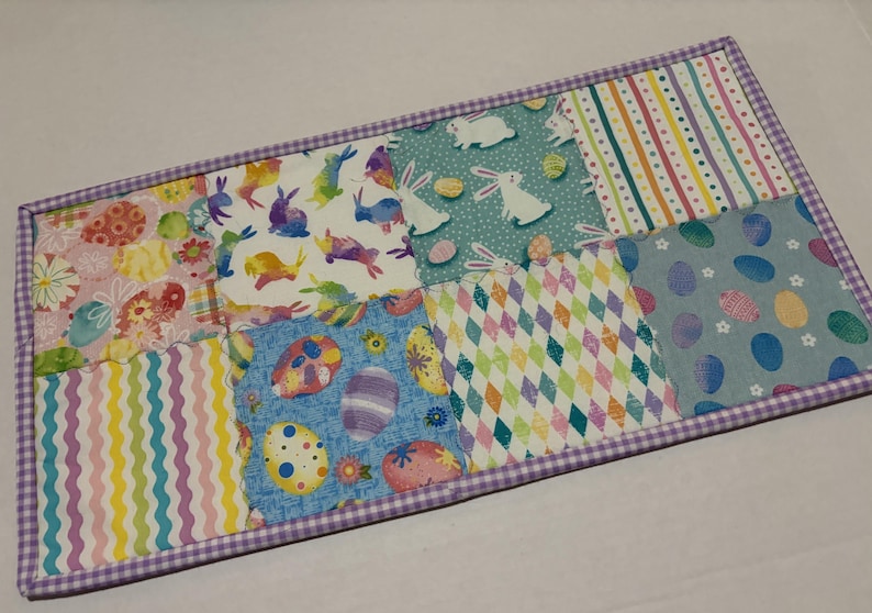 Może przedstawiać: Prostokątna, pikowana podkładka pod talerz w tematyce wielkanocnej. Podkładka ma wz&oacute;r patchworkowy z r&oacute;żnymi wzorami, w tym pisankami, kr&oacute;liczkami i kolorowymi kształtami geometrycznymi. Krawędzie obszyte są fioletowo-białą tkaniną w kratkę.