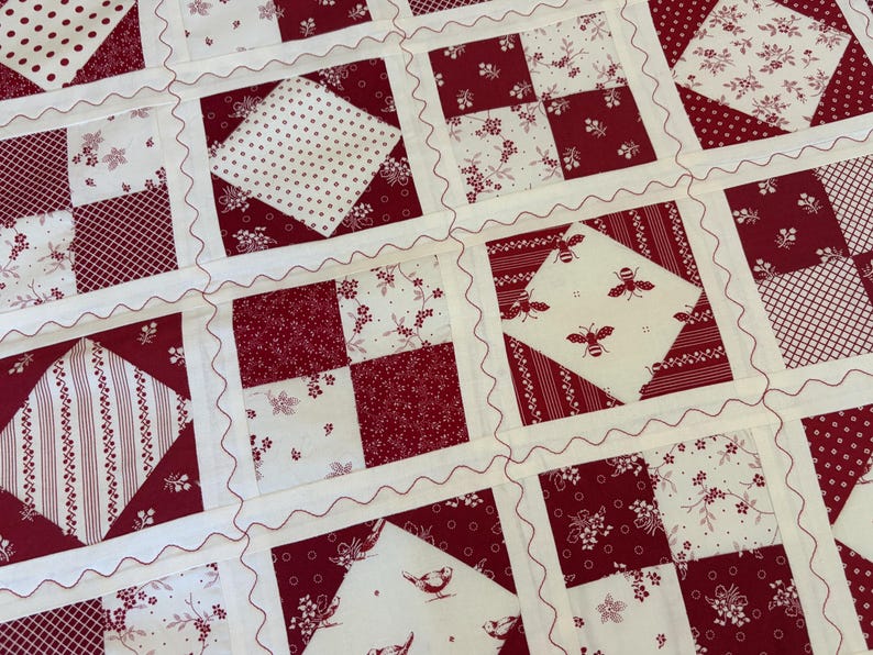 May include: A patchwork quilt featuring a repeating grid of squares in deep red and white. The design includes various patterns such as floral prints, polka dots, stripes, and small bee and bird motifs, all separated by white sashing with wavy stitching.