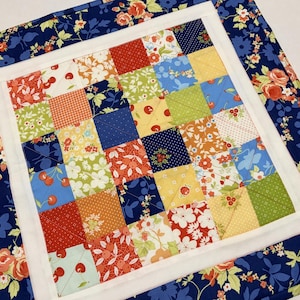 May include: A colorful patchwork quilt with a square pattern. The center features small squares in various patterns and colors, including red, green, yellow, and blue. A white border surrounds the center, and a floral patterned blue fabric frames the quilt.