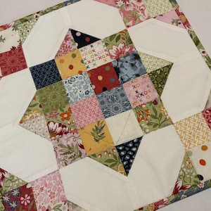 May include: A square quilted fabric piece featuring a central star motif composed of multi-colored patchwork squares in shades of pink, yellow, green, blue, and white. The design includes floral, polka dot, and geometric patterns with white fabric accents.