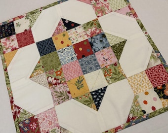 May include: A square quilted fabric piece featuring a central star motif composed of multi-colored patchwork squares in shades of pink, yellow, green, blue, and white. The design includes floral, polka dot, and geometric patterns with white fabric accents.