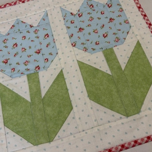 May include: A quilted placemat featuring two tulip designs. The tulip heads are light blue with a floral pattern, and the stems and leaves are green. The placemat has a white background with small blue dots and a red and white gingham border.