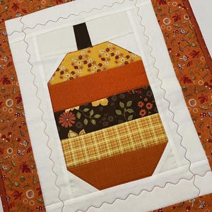 May include: A quilted wall hanging with a patchwork design of an orange, brown, and yellow acorn. The acorn is surrounded by a white border with a scalloped edge. The background is a brown fabric with a small orange and brown floral pattern.