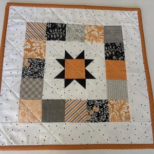 May include: A square quilted fabric mat featuring a central orange star surrounded by a border of alternating black, white, and orange patterned fabric squares. The outer border is white with small black and orange dots and an orange binding.
