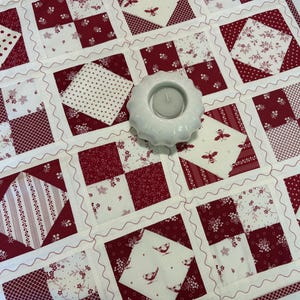 May include: A square patchwork quilt featuring a repeating grid of red and white fabric squares with various patterns including polka dots, floral prints, bees, and birds, accented by wavy white stitching and a white ceramic tea light holder.