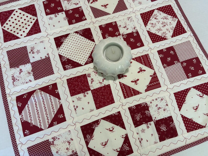 May include: A square patchwork quilt featuring a repeating grid of red and white fabric squares with various patterns including polka dots, floral prints, bees, and birds, accented by wavy white stitching and a white ceramic tea light holder.